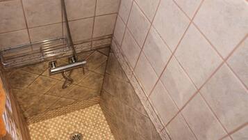 Un baño pequeño con azulejos, zona de ducha, un estante metálico y desagüe.