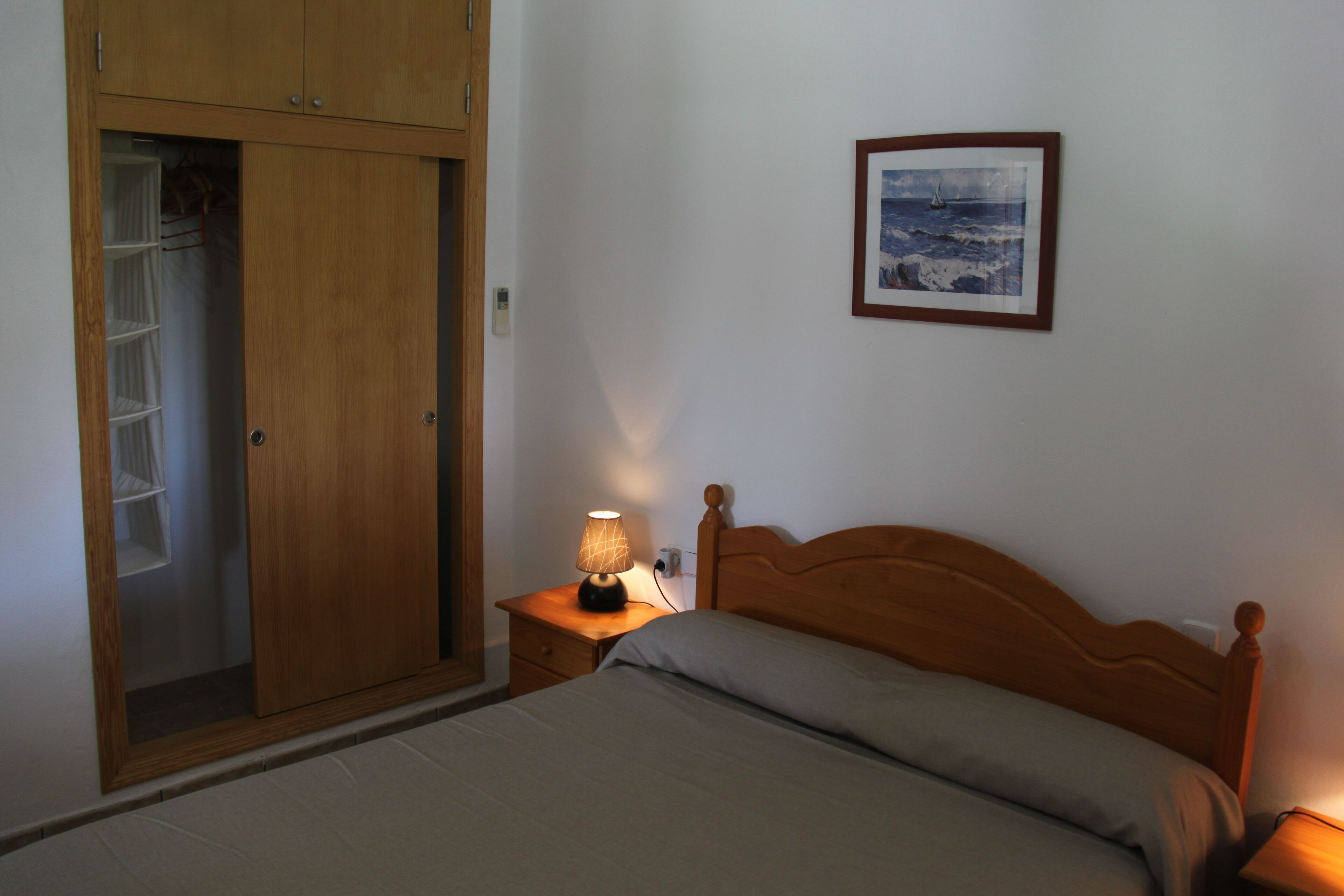 Ein Schlafzimmer mit einem Bett, einem Nachttisch mit Lampe, einem Holzschrank und einem gerahmten Bild an der Wand.