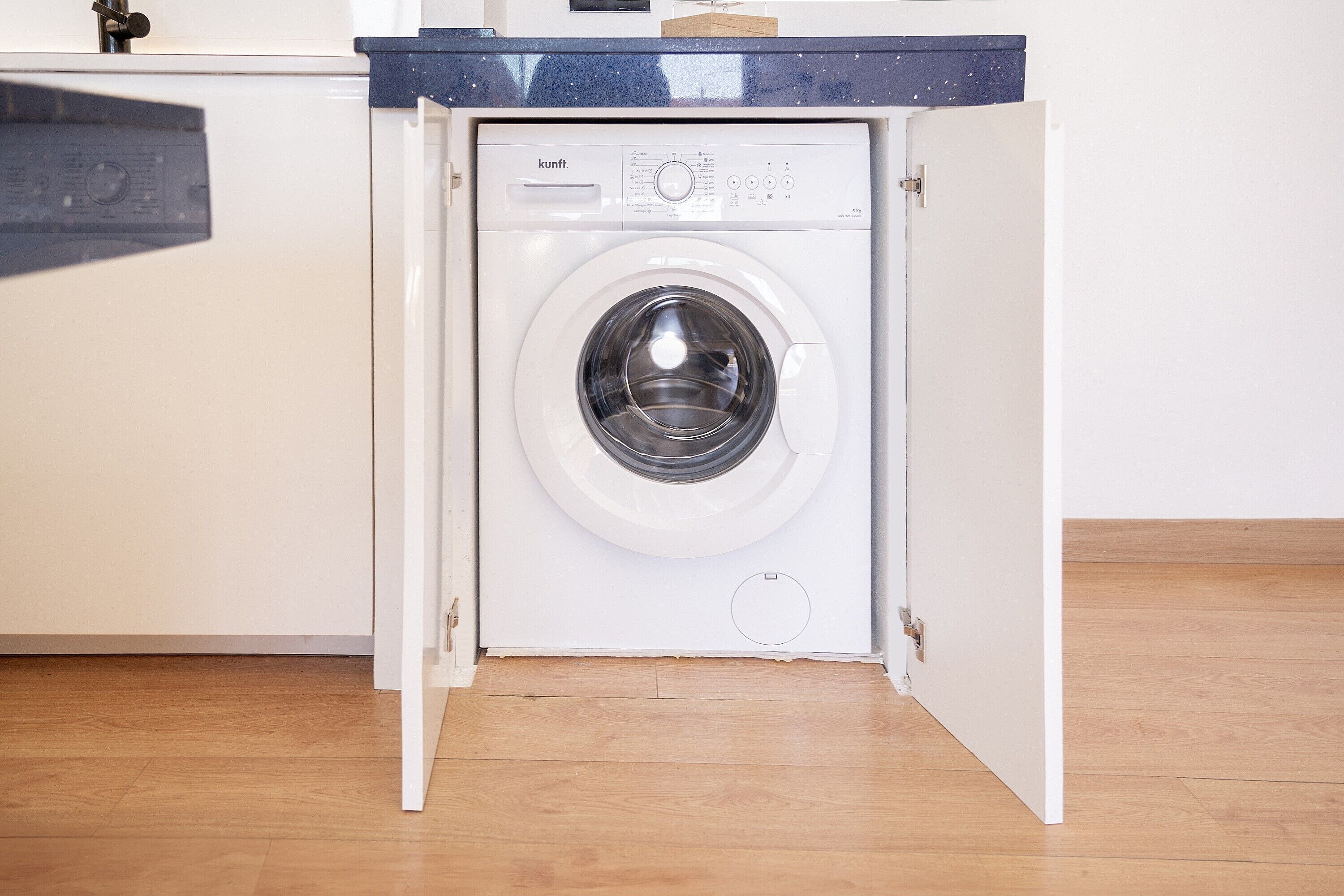 A compact, white front-loading washing machine with a control panel and a door, installed under a kitchen counter with a dark countertop.