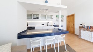 Een moderne keuken met een blauw aanrecht, witte kasten en een houten deur. Voor het aanrecht staan drie witte stoelen.