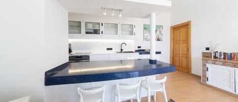 Una cocina moderna con encimera azul, armarios blancos y una puerta de madera. Hay tres sillas blancas frente al mostrador.