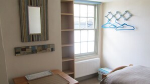 Un dormitorio con una cama de madera, una mesita con un tapete, un radiador y una ventana que da a la calle.