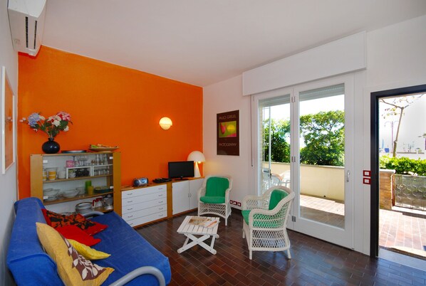 Une pièce comprenant un canapé bleu, un fauteuil en osier, une petite table, une étagère avec des ustensiles de cuisine, une lampe fixée au mur et une porte-fenêtre coulissante donnant sur un espace extérieur.