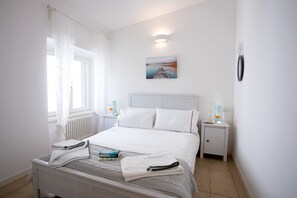 Ein ordentlich bezogenes Bett mit weißen Laken, ein Nachttisch mit Lampe und ein Bild an der Wand.