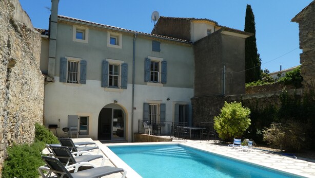 Un espace aménagé au bord de la piscine, avec des chaises longues, une piscine et un bâtiment aux murs en pierre et aux volets bleus.