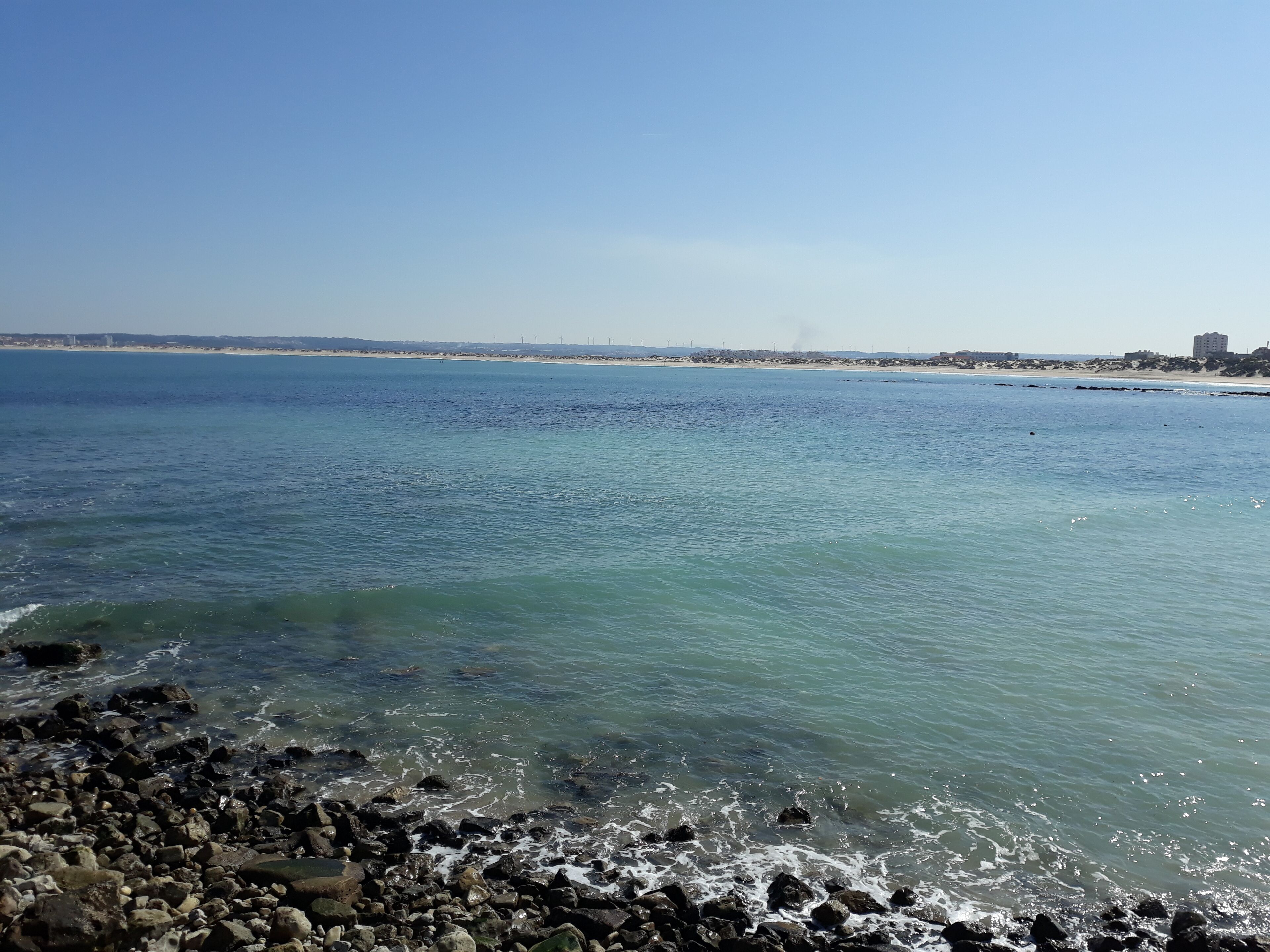 Une vue sur la côte, avec une eau cristalline, un rivage rocheux et des bâtiments qui se dessinent à l’horizon.