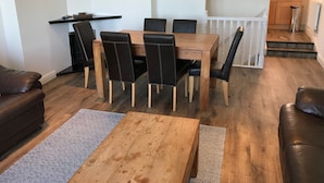 A modern living room with a wooden dining table and black chairs, a brown leather sofa, and a kitchen in the background.
