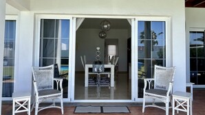 Ein überdachter Patio-Bereich mit zwei Korbstühlen, einem Esstisch mit Stühlen und einer Glasschiebetür zu einem Innenraum mit einer Kronleuchter.