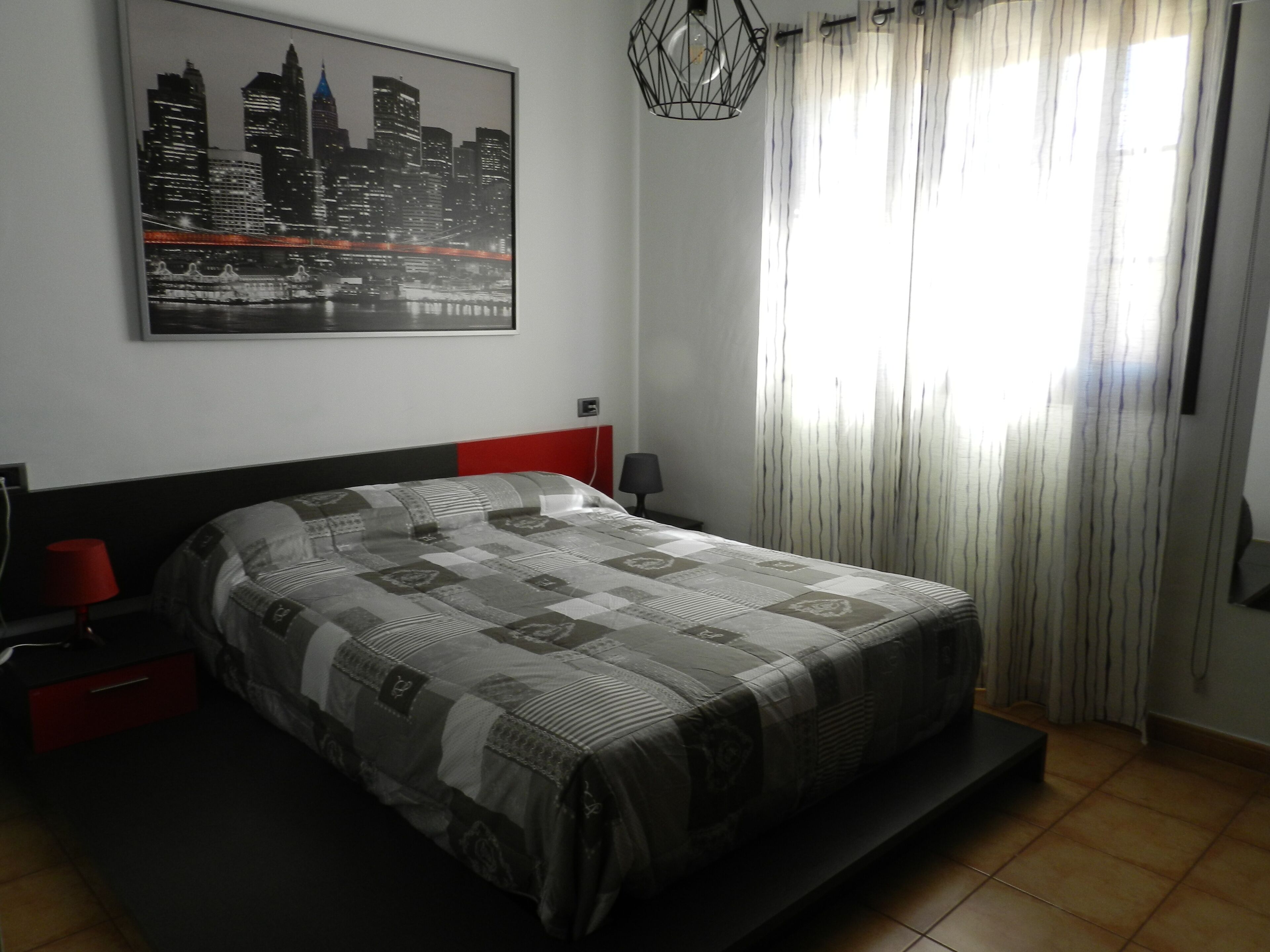 A bedroom with a bed, a red nightstand, a lamp, a cityscape painting, and a window with curtains.