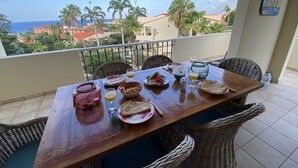 Un balcon avec une table en bois mise pour le petit-déjeuner, donnant sur une zone côtière bordée de palmiers et de bâtiments.