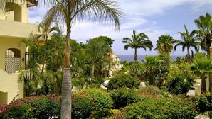 Un jardin luxuriant, agrémenté de palmiers et de plantes à fleurs, un bâtiment aux fenêtres en arc de cercle et une vue sur la mer.