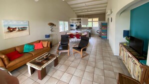Un vaste espace de vie comprenant un canapé, des fauteuils, une table basse et un téléviseur. On y trouve également un ventilateur de plafond, un tableau représentant des bateaux et, au fond, une cuisine.