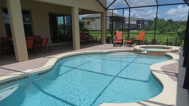A swimming pool with a tiled deck, surrounded by chairs and a covered patio area.