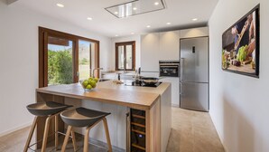 A modern kitchen with a central island, bar stools, a refrigerator, and a window overlooking greenery. - Villa Can Dala luxury in Port de Pollensa Sleeps 12 (Puerto Pollensa)