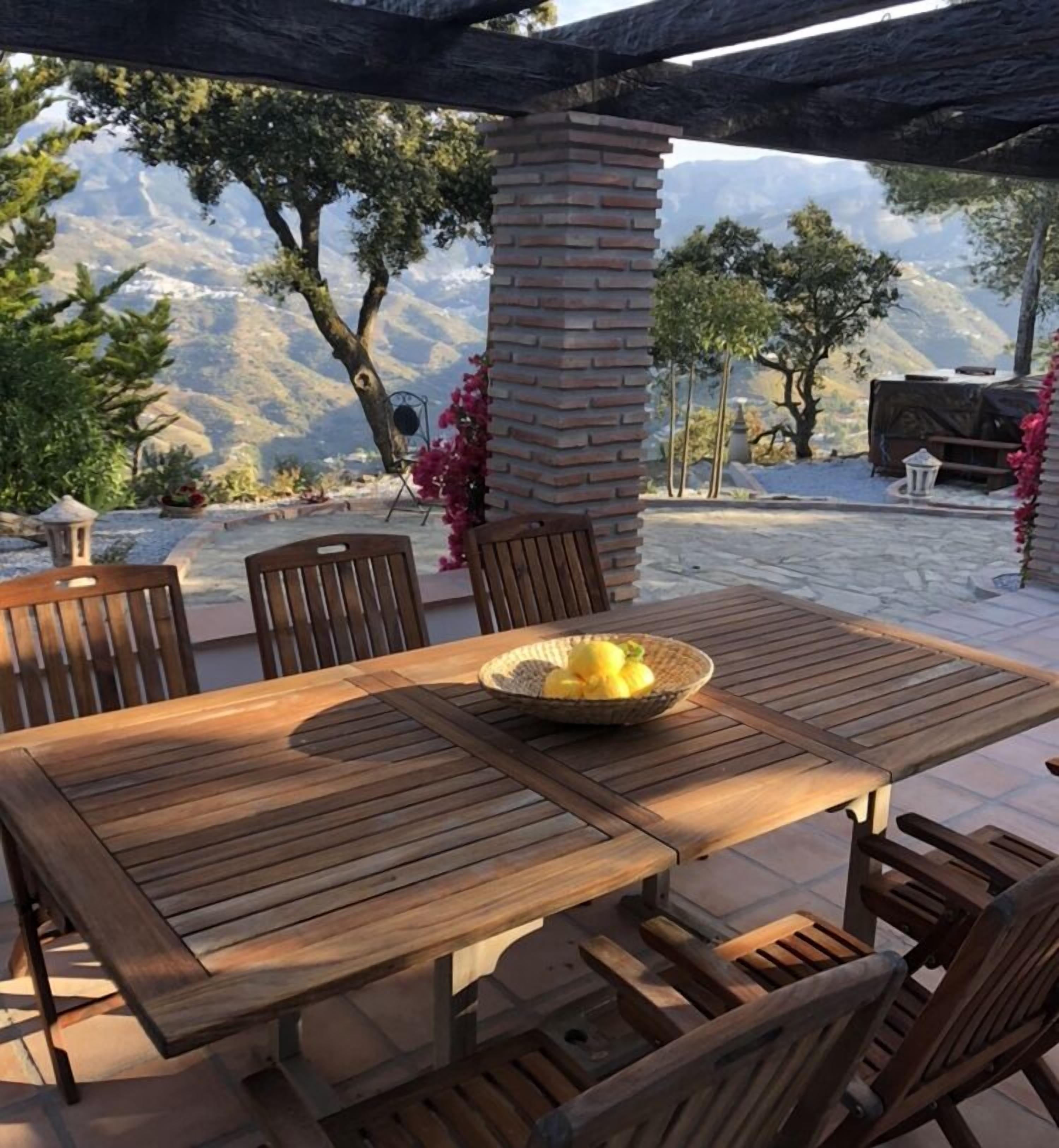 An outdoor patio with a wooden table and chairs, a brick pillar, and a view of mountains.