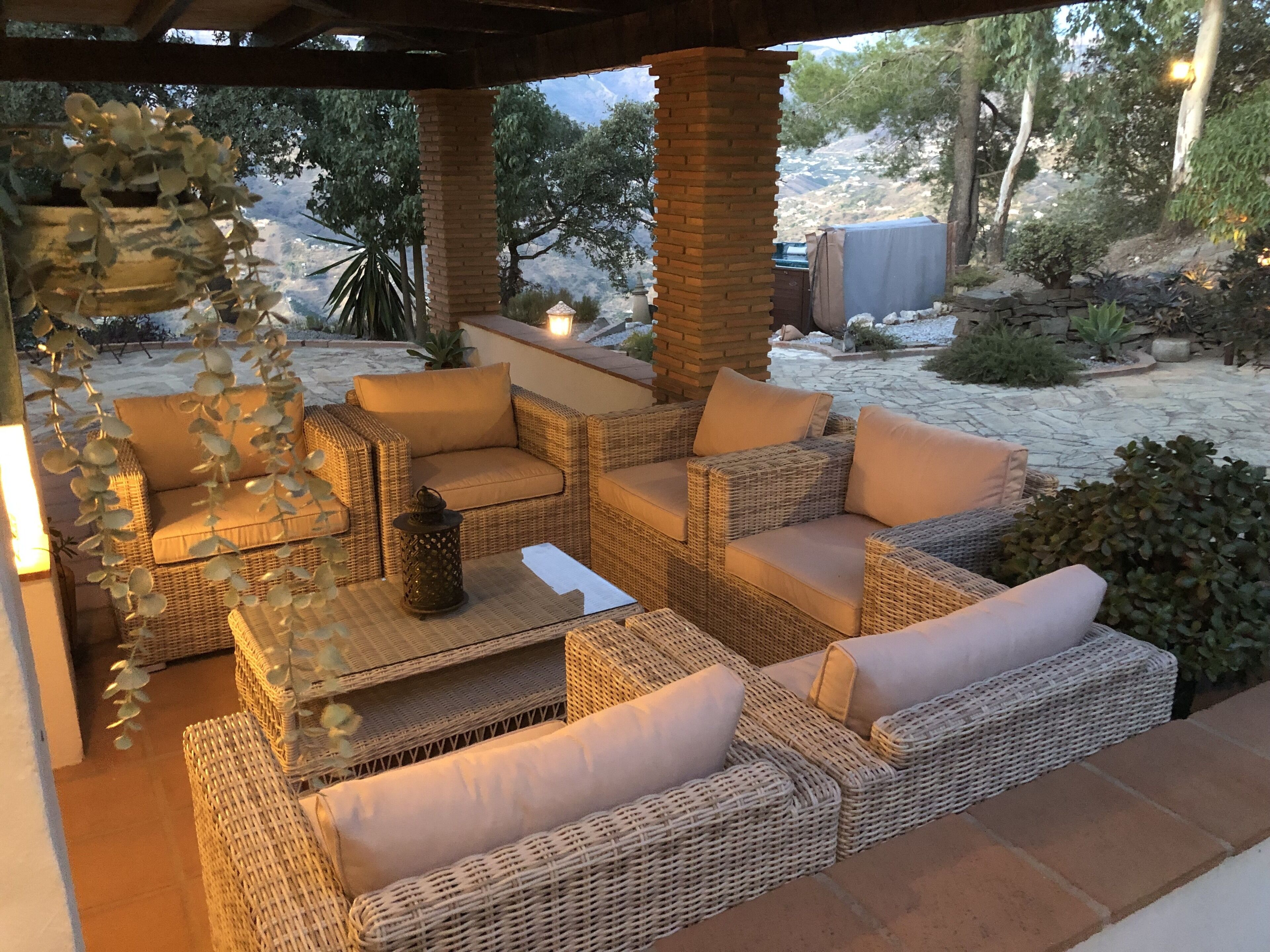A covered outdoor seating area with wicker furniture and cushions, surrounded by stone walls and plants.