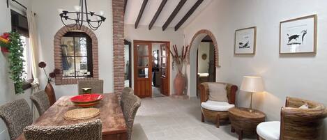A dining area with a wooden table and wicker chairs, a chandelier, and a view of the kitchen through an archway.
