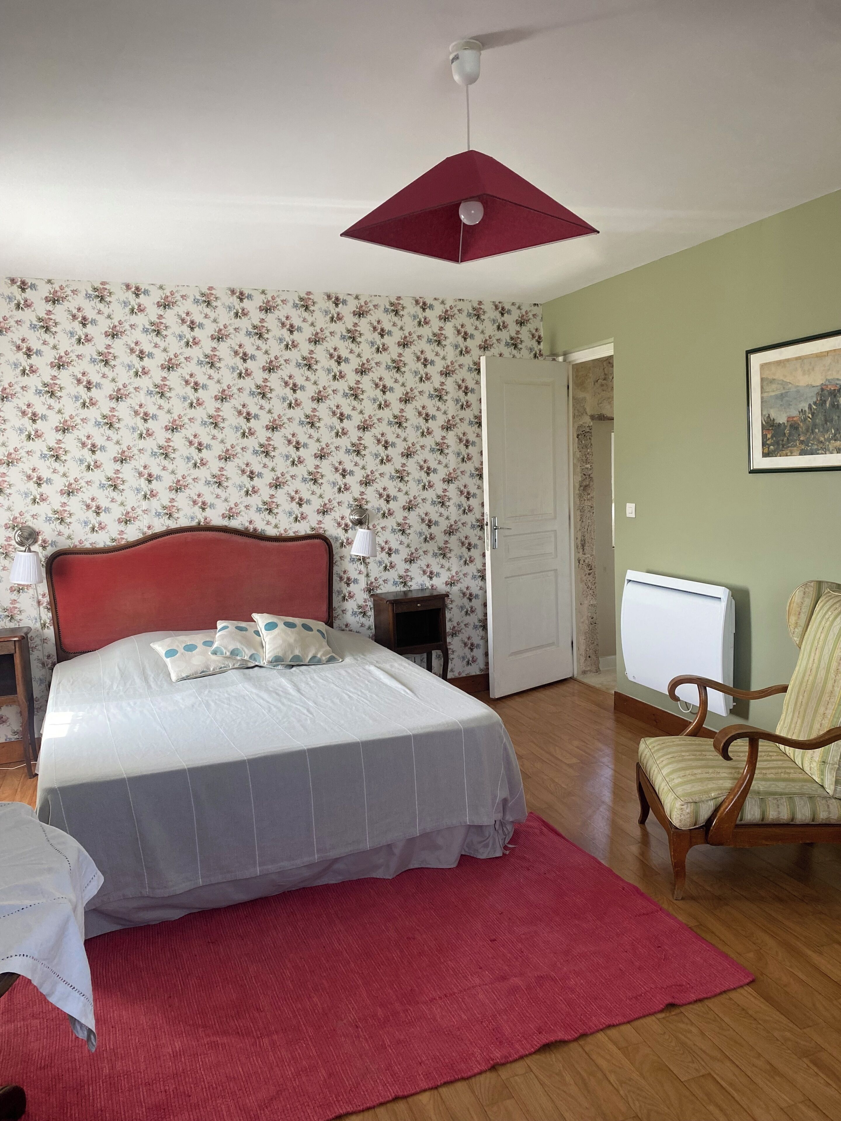 A bedroom with a bed, a red headboard, a nightstand with a lamp, a chair, a radiator, and a door.