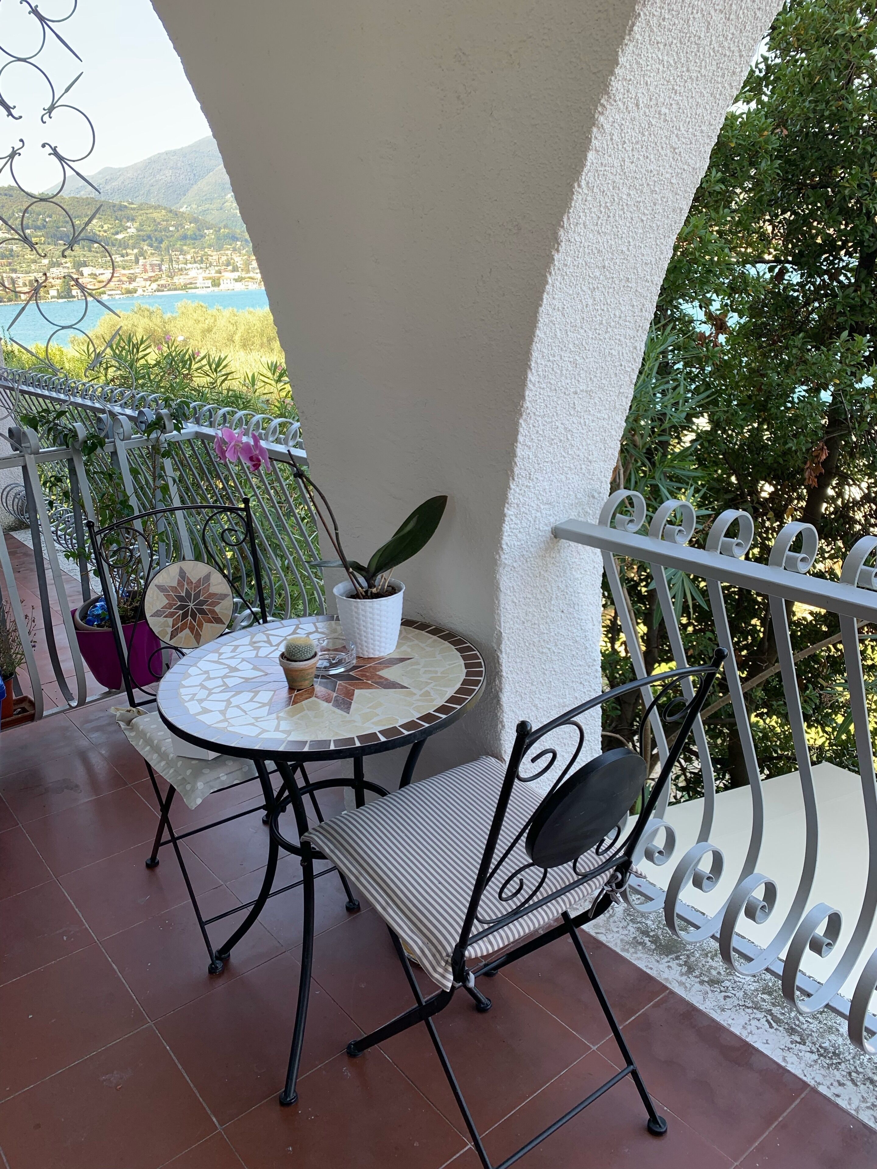 A small outdoor patio with a round table and a chair, surrounded by metal railings and potted plants.