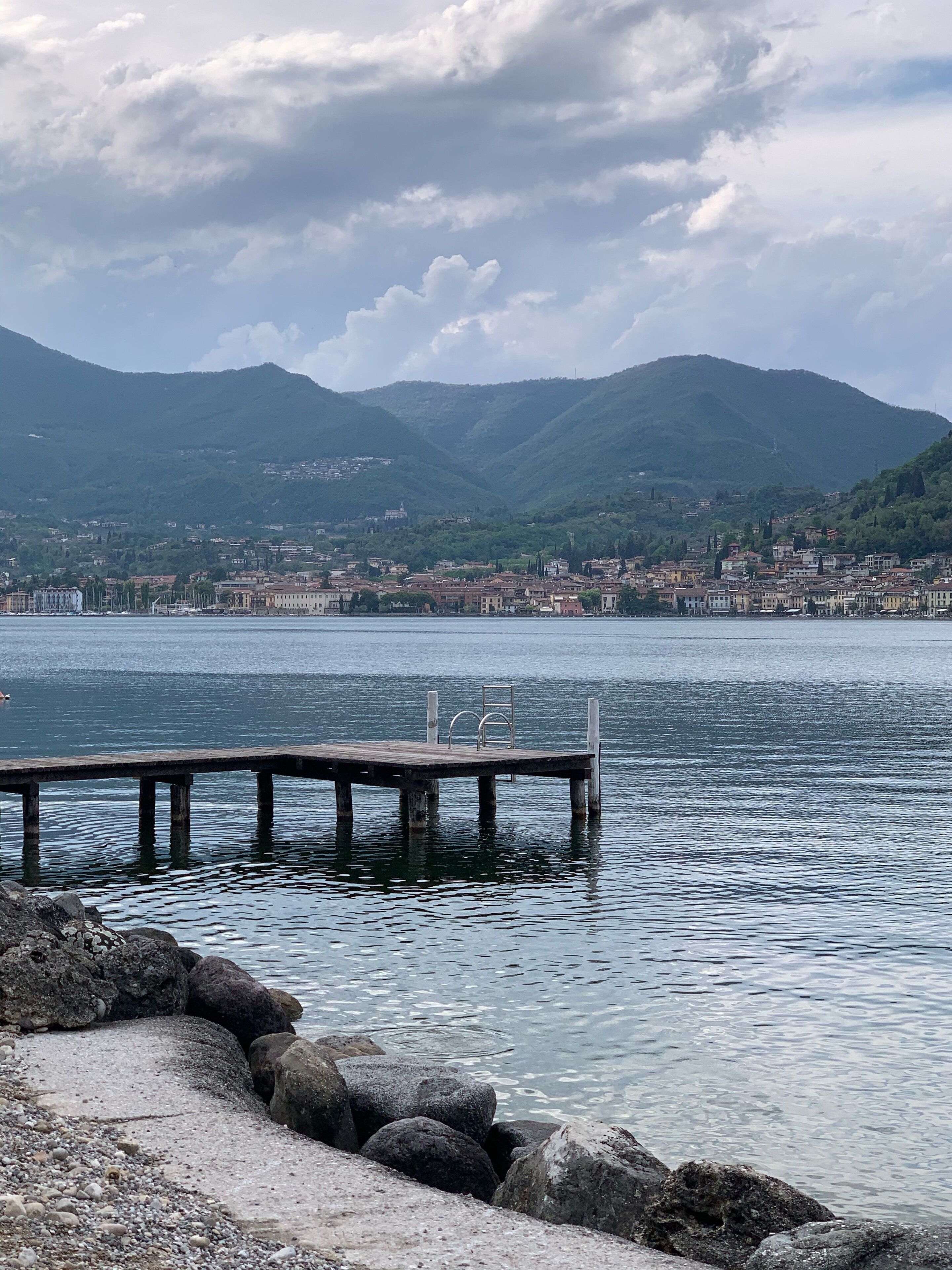 Vista a un lago con un muelle que se adentra en aguas tranquilas, rodeado de terreno rocoso y un paisaje urbano distante con montañas al fondo.