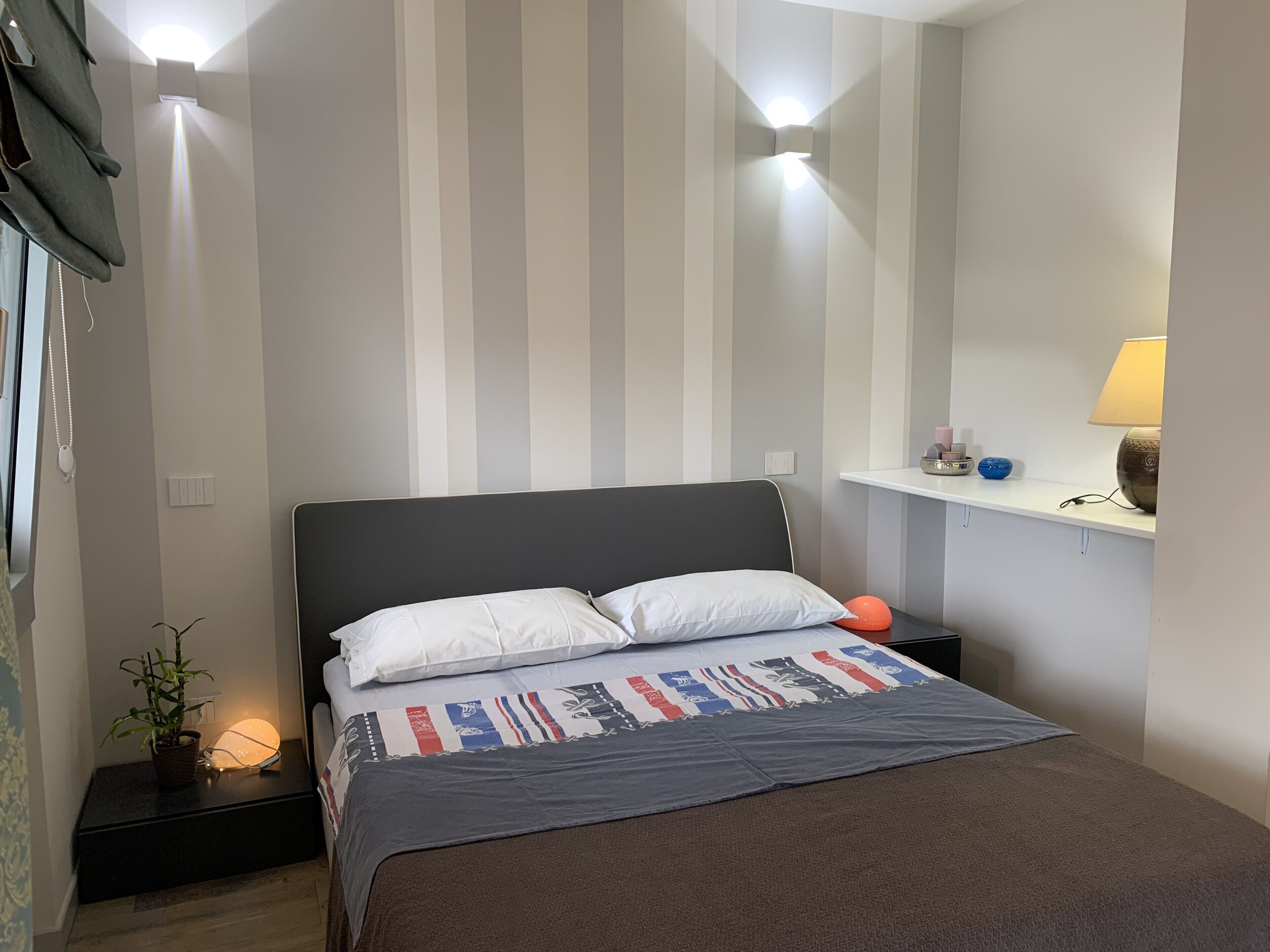 Un lit bien fait, recouvert d’une couvre-lit à motifs, une lampe de chevet et une petite table de nuit agrémentée d’une plante.