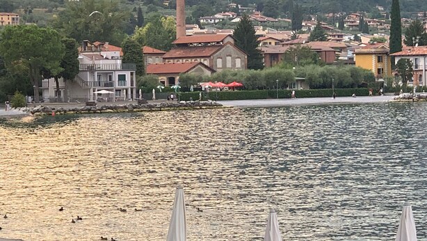 Vista del lago con una fila de sillas reclinables azules y sombrillas blancas, una orilla rocosa y edificios al fondo.