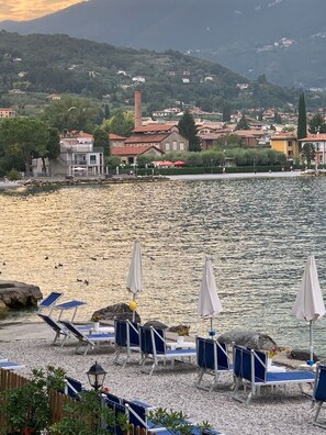 Une vue sur le lac, avec une rangée de chaises longues bleues et de parasols blancs, une rive rocheuse et des bâtiments en arrière-plan.