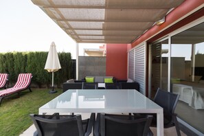 An outdoor patio with a glass-top table, black chairs, striped lounge chairs, a white umbrella, and a sofa with green cushions.