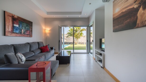 Ein modernes Wohnzimmer mit einem grauen Sofa, einem roten Beistelltisch und einem Fernseher auf einem Lowboard. An der Wand hängt ein großes Gemälde und durch die Glasdoor ist ein Pool und Palmen zu sehen.
