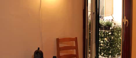 Quarto nos fundos com uma cadeira e uma mesinha, porta de vidro deslizante para uma varanda e luminária fixada na parede.