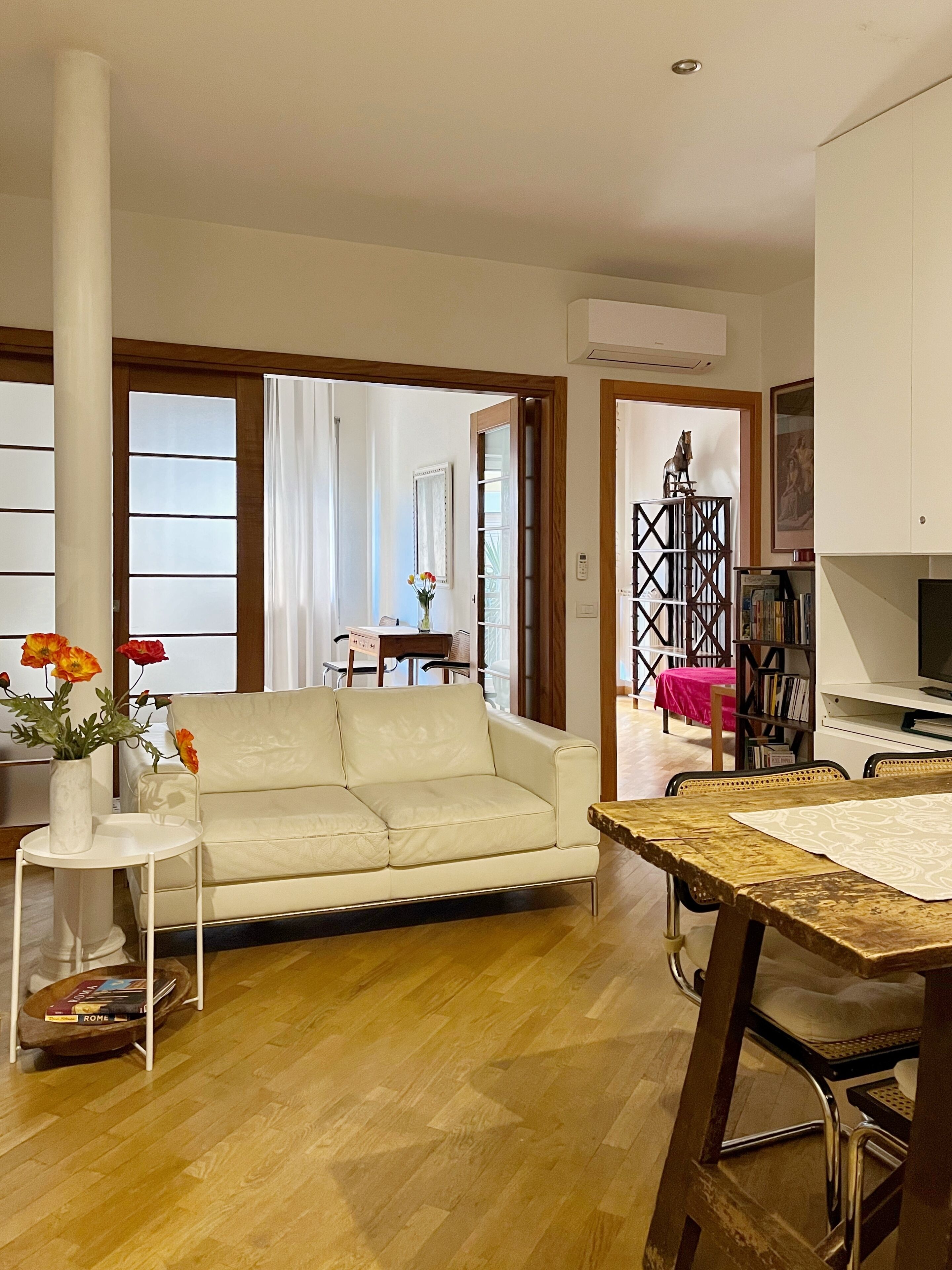 A living room with a white sofa, a small round table with a vase of flowers, and a wooden dining table with chairs.