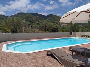 A rectangular swimming pool with a tiled edge, surrounded by a brick patio. There are two wicker lounge chairs and a small table under a white umbrella. The background features a mountainous landscape with greenery and a few buildings. - Luxury  Villa, panoramic sea view, peaceful surroundings, near Nafpaktos city (Ano Platanitis Nafpaktos)