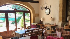 Una cocina rústica con una isla de madera, una mesa de comedor y sillas. Se aprecia un gran ventanal con arco, una araña y un reloj en la pared.