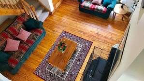 A living room with a wooden floor, a green sofa with patterned cushions, a wooden coffee table with a vase of flowers, and a window with curtains.