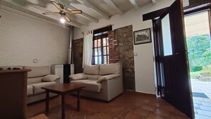 A living room with a sofa, a coffee table, a ceiling fan, a fireplace, and a door leading outside. - Oruna De Pielagos: Country Cottage / Gite - Santander (Oruña de Piélagos)