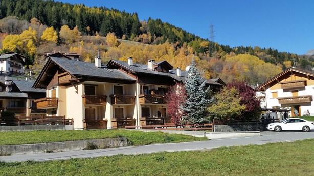 A row of traditional wooden buildings with balconies, set against a backdrop of autumn-colored trees and a clear blue sky.