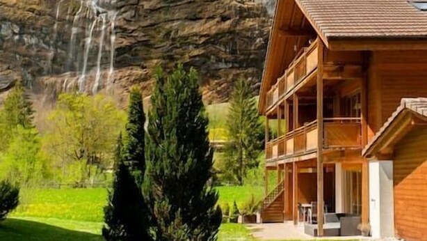 A wooden chalet with a balcony, a waterfall in the background, and a forested mountain. - Beautiful apartment with balcony and waterfall view (Lauterbrunnen)