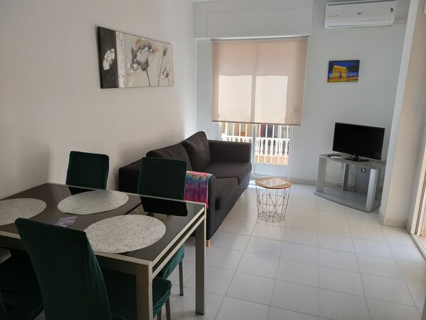 A living room with a sofa, a dining table with chairs, a TV on a stand, and artwork on the walls.