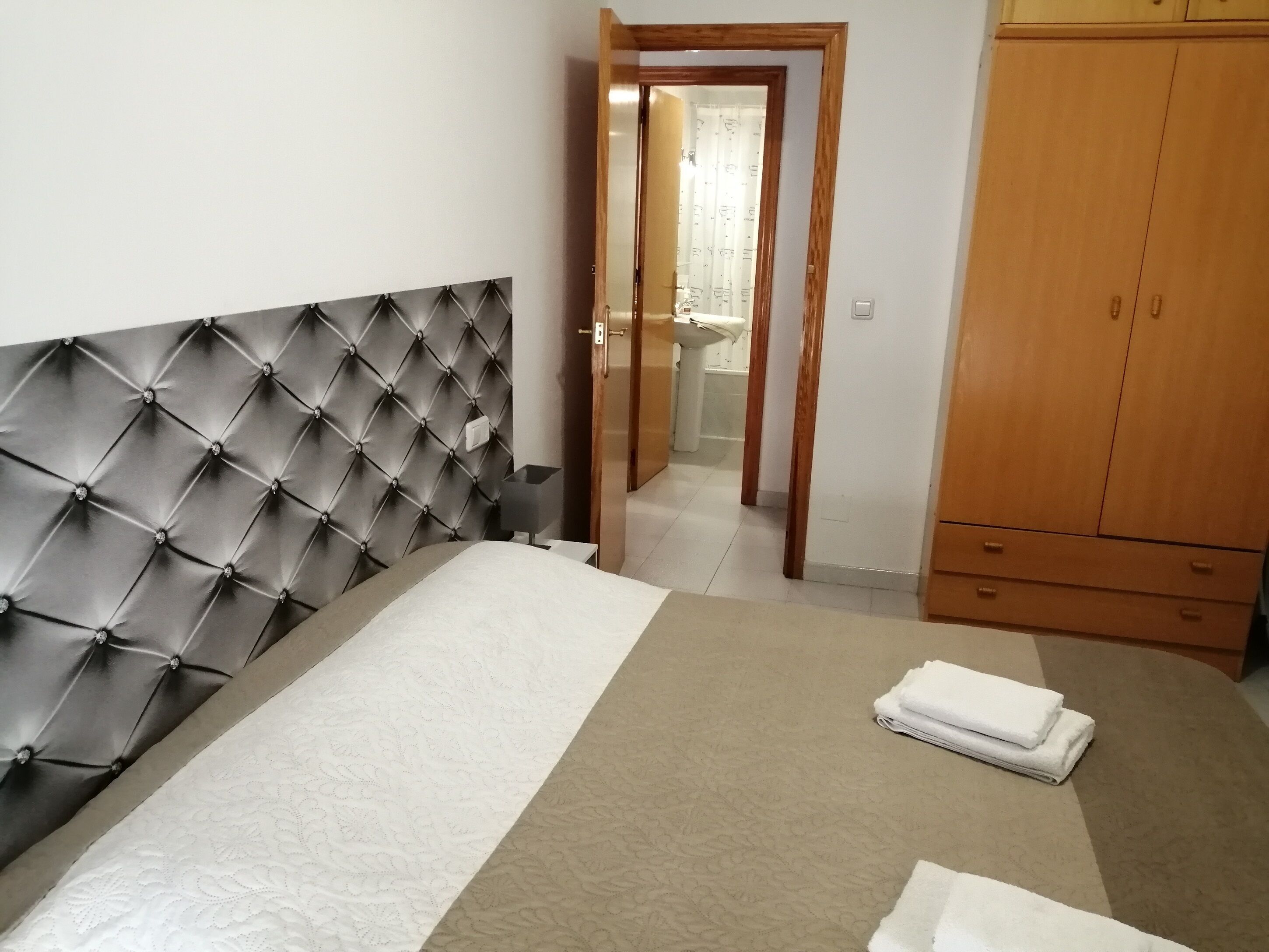 Une chambre à coucher avec une tête de lit capitonnée, un lit recouvert d’une couvre-lit à motifs, une armoire en bois et une salle de bain visible à travers une porte ouverte.