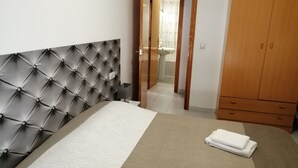 A bedroom with a tufted headboard, a bed with a patterned bedspread, a wooden wardrobe, and a bathroom visible through an open door. - Holiday Houses - Playa del Cura 2 (Torrevieja)