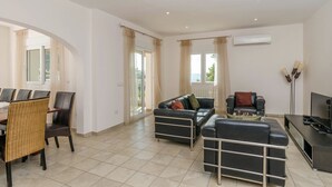 En rummelig opholdsstue med spiseplads, sofa, TV-møbel og et stort vindue med gardiner.