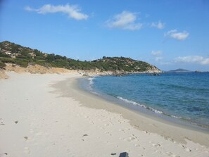 En sandstrand med klart, blåt vand, klippeudspring og en bakke med bygninger.
