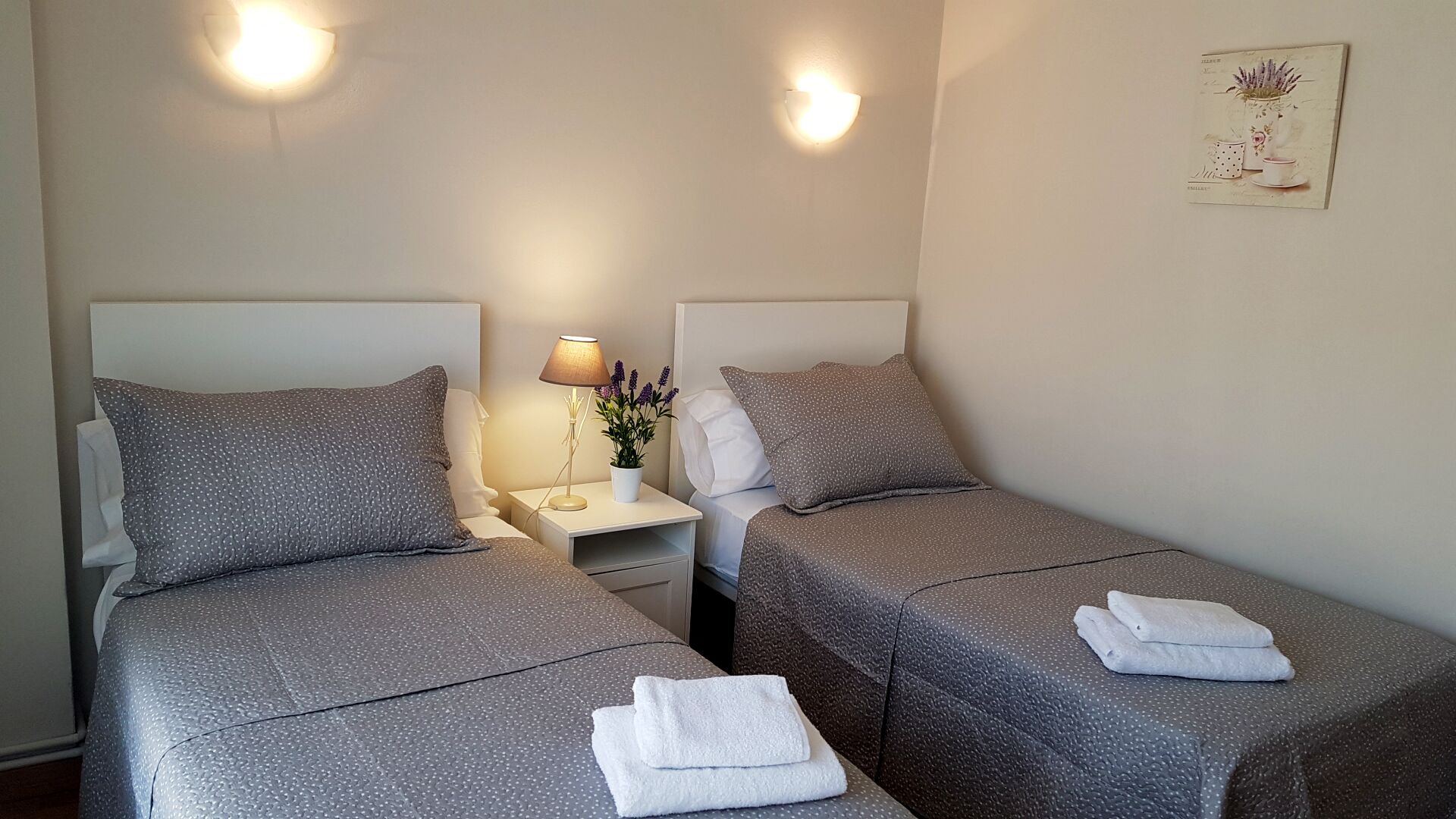 A twin room with two beds, a nightstand with a lamp, and a wall-mounted light fixture.