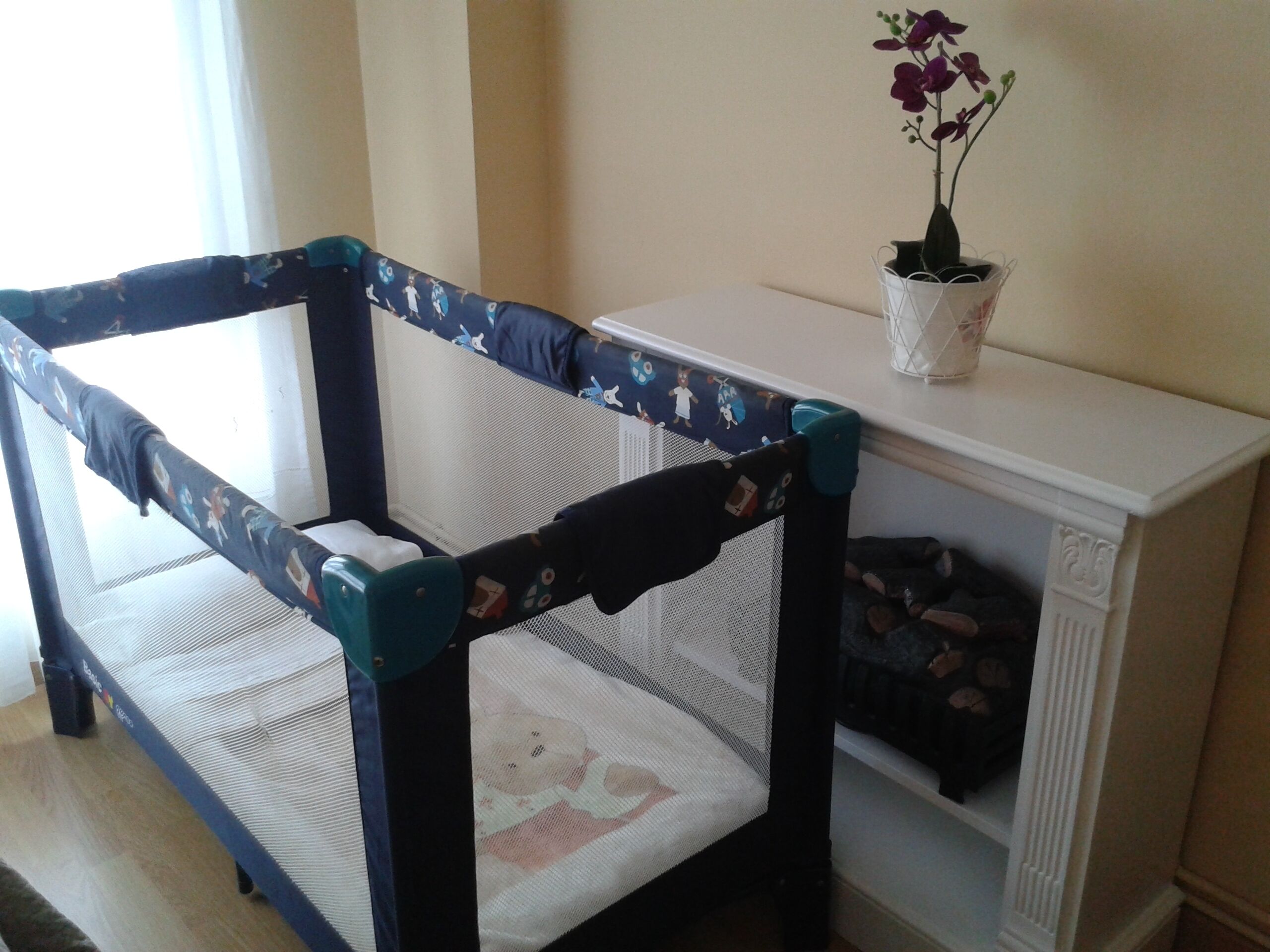 A baby crib with a mesh and padded mattress, positioned next to a white shelf with a potted plant.