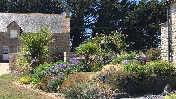 Un bâtiment en pierre doté d’un petit porche et d’un jardin où poussent différentes plantes et fleurs.