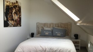 Une chambre avec un plafond en pente, un lit avec une literie blanche, deux oreillers ornés du mot « OCÉAN », un tableau au mur et une petite table de chevet avec une lampe.