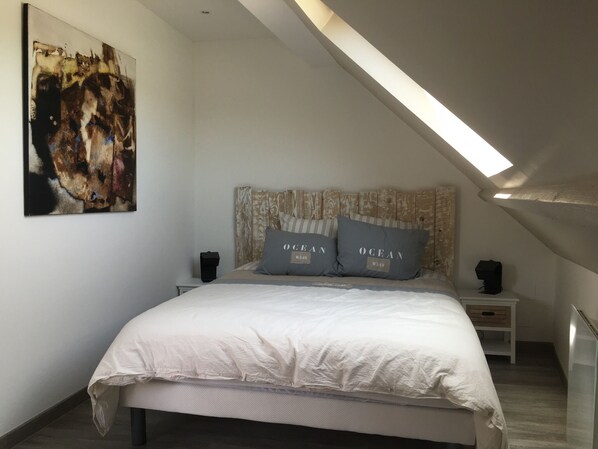 A bedroom with a sloped ceiling, a bed with white bedding, two pillows with 'OCEAN' text, a painting on the wall, and a small bedside table with a lamp. - Cozy cottage 700m from the beach (Plouhinec)