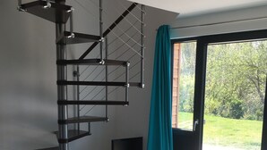 Un salon moderne avec un escalier en colimaçon, un fauteuil confortable, une table basse en bois et une grande fenêtre donnant sur un paysage verdoyant.