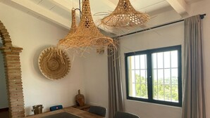 A conference room with a wooden table, two woven pendant lights, a brick column, a window with curtains, and a view of greenery outside.