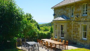 A stone building with a large patio table and chairs, surrounded by lush greenery and a view of the landscape. - MANOR ON COAST WITH SEA VIEWS, TENNIS COURT, BIG GAMES ROOM + BAR, GARDENS (Ventnor)